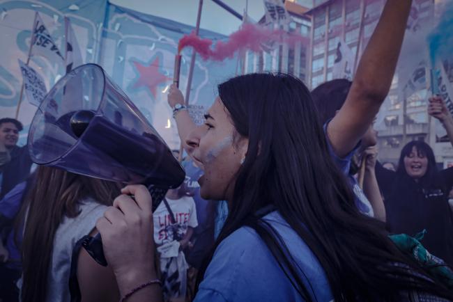 Un manifestante sobre la Avenida 9 de Julio sostiene un altavoz y grita lemas en defensa de la educación pública. Detrás de elle, otres portan bengalas azules y rojas. Buenos Aires. 23 de abril de 2024. (Lizbeth Hernández)