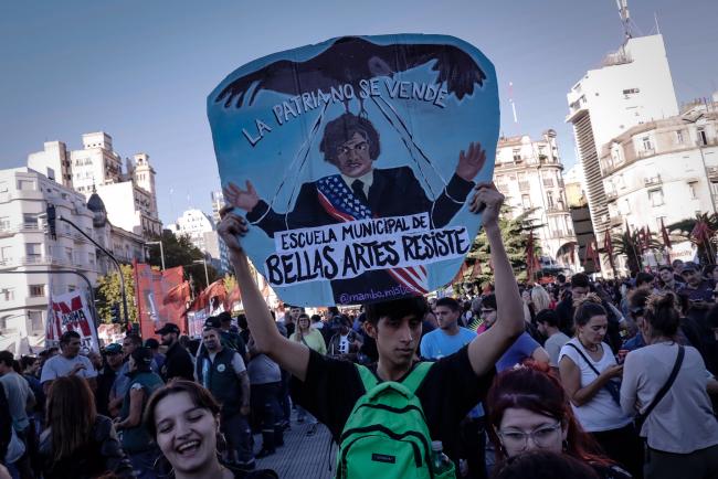 One student holds a poster created by another student (IG: mambo.mistico). Milei is depicted as a puppet of the United States, and a bald eagle pulls his strings. Buenos Aires. April 23, 2024. (Lizbeth Hernández)