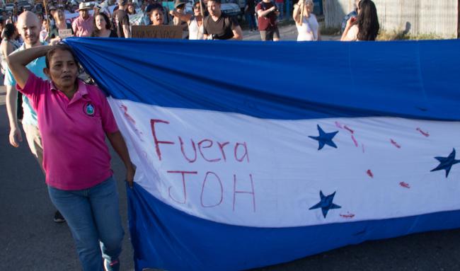 International accompaniers join Hondurans marching with a flag reading "Fuera JOH" (Out with JOH), short for President Juan Orlando Hernández. (Peg Hunter / Flickr / CC BY-NC 2.0)
