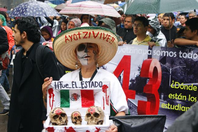 A protester holds a sign that reads “Fue El Estado” (It was the state), with the Mexican flag and three bloody skulls, at the first-anniversary march commemorating the 43 who disappeared at Ayotzinapa. (PetrohsW/Wikimedia Commons/CC BY-SA 4.0) 