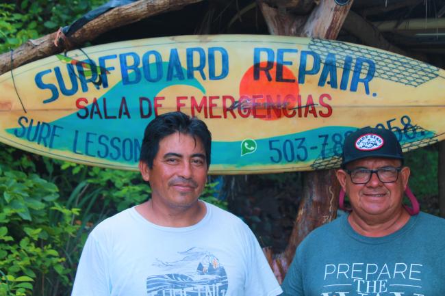 Turcios and Yepi posing under the sign of their surfboard repair shop on Las Flores beach. (Julián Reingold)