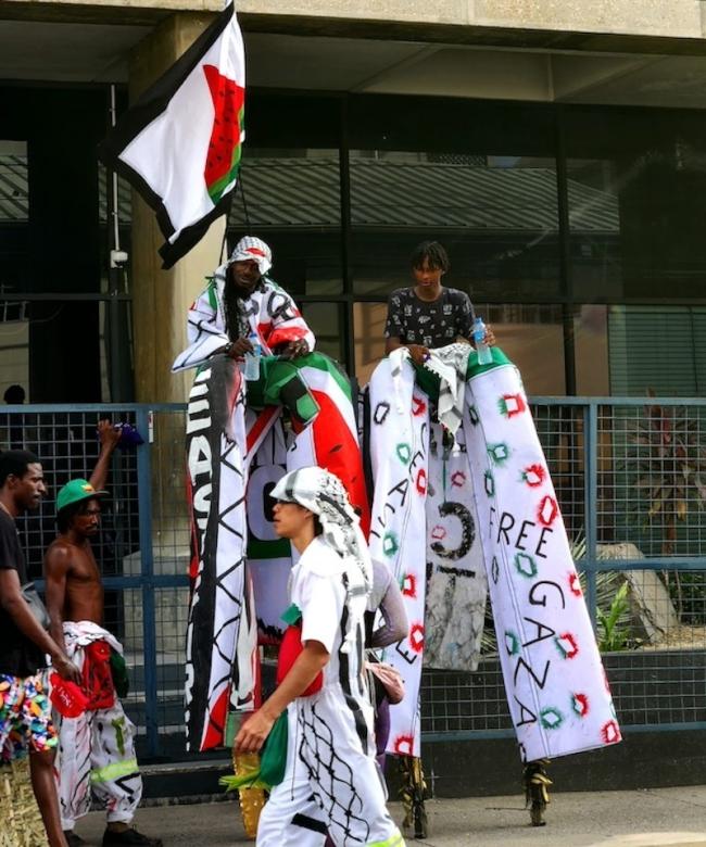 Vulgar Fraction's 2024 presentation, "Isabày: Bear With-ness," which aimed to display solidarity with Palestine. Two "moko jumbies" (stilt walkers) are seated while two regular masqueraders and a member of the public walk by. (Khalea Robertson)